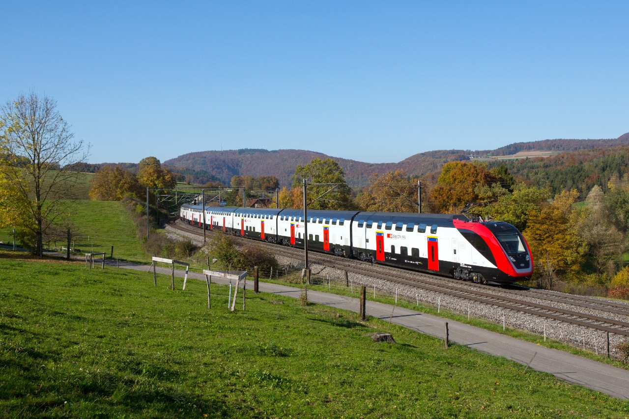 FV-Dosto fährt an einem sonnigen Tag in Zeihen auf der Bözbergstrecke zwischen Basel und Brugg. Links vom Zug befindet sich eine Wiese mit einem kleinen Spazierweg und rechts ist ein Wald in Herbstfarben.