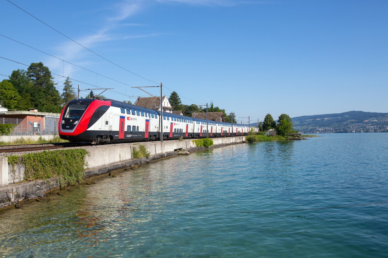 Der FV-Dosto fährt an einem sonnigen Tag am Zürichsee in Richterswil vorbei. Der See befindet sich auf der linken Seite des Zuges.