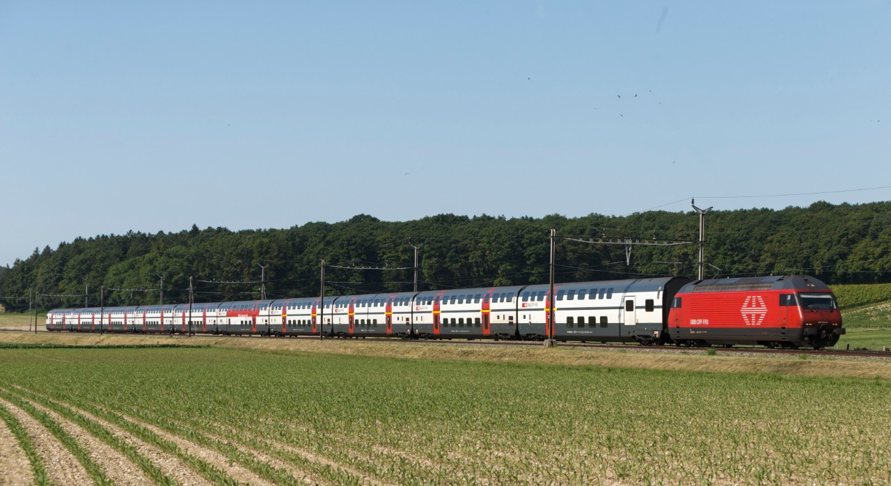 IC2000: Aussenansicht des IC2000. Der Zug, bestehend aus 10 Wagen und einer Lokomotive, fährt bei sommerlichem Wetter durch die Landschaft. Im Hinergrund befindet sich ein Wald. Am Himmel fliegen Vögel ummher.